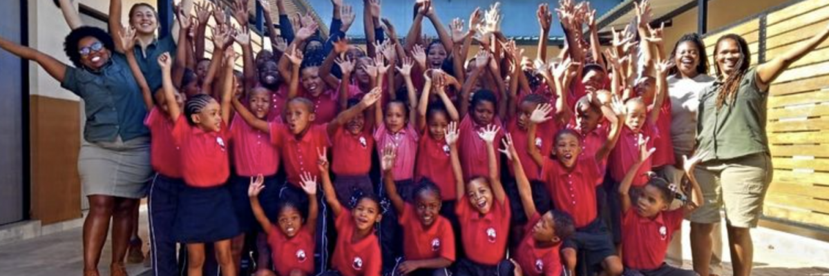 Groupe d’enfants San et enseignants levant les bras et souriant dans la cour de leur école primaire en Namibie, symbole de joie et de réussite collective.