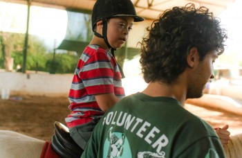 Un volontaire soutenant un enfant portant un casque de protection lors d'une séance de thérapie équine.