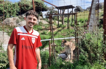 Un volontaire de Globalong souriant, vêtu d'un maillot rouge, pose devant l'enclos d'un puma dans une réserve naturelle verdoyante.
