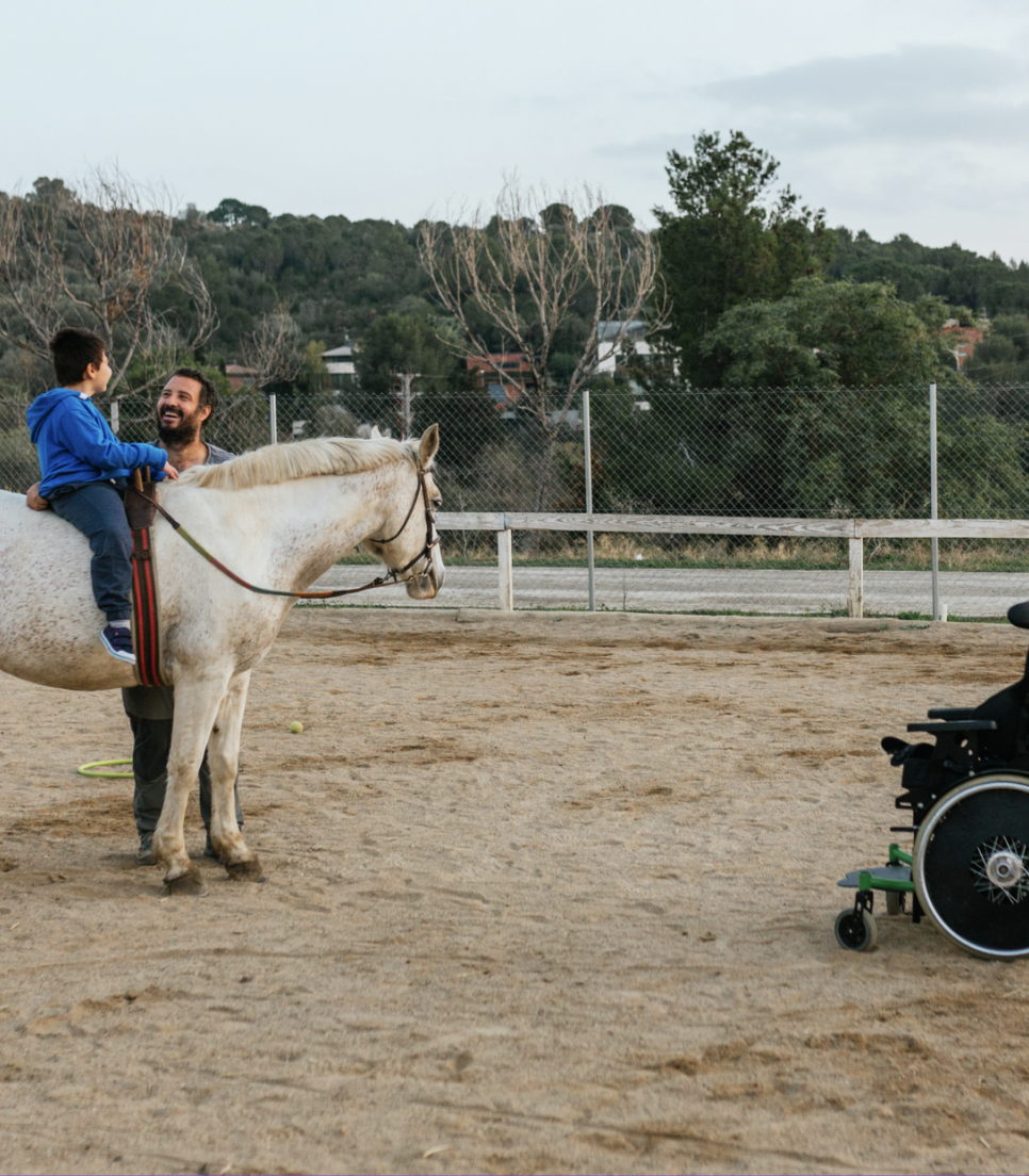 Un enfant assis sur un cheval lors d'une séance de thérapie équine, avec un bénévole souriant à ses côtés et un fauteuil roulant visible en arrière-plan, dans le cadre du programme Globalong.