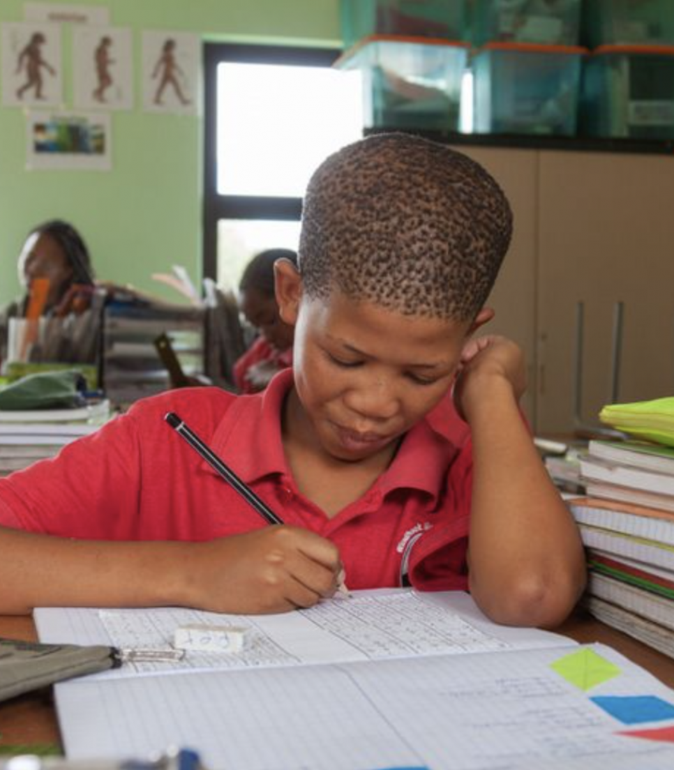 Élève San concentré sur un exercice d’écriture dans une salle de classe en Namibie, entouré de cahiers et de matériel scolaire.