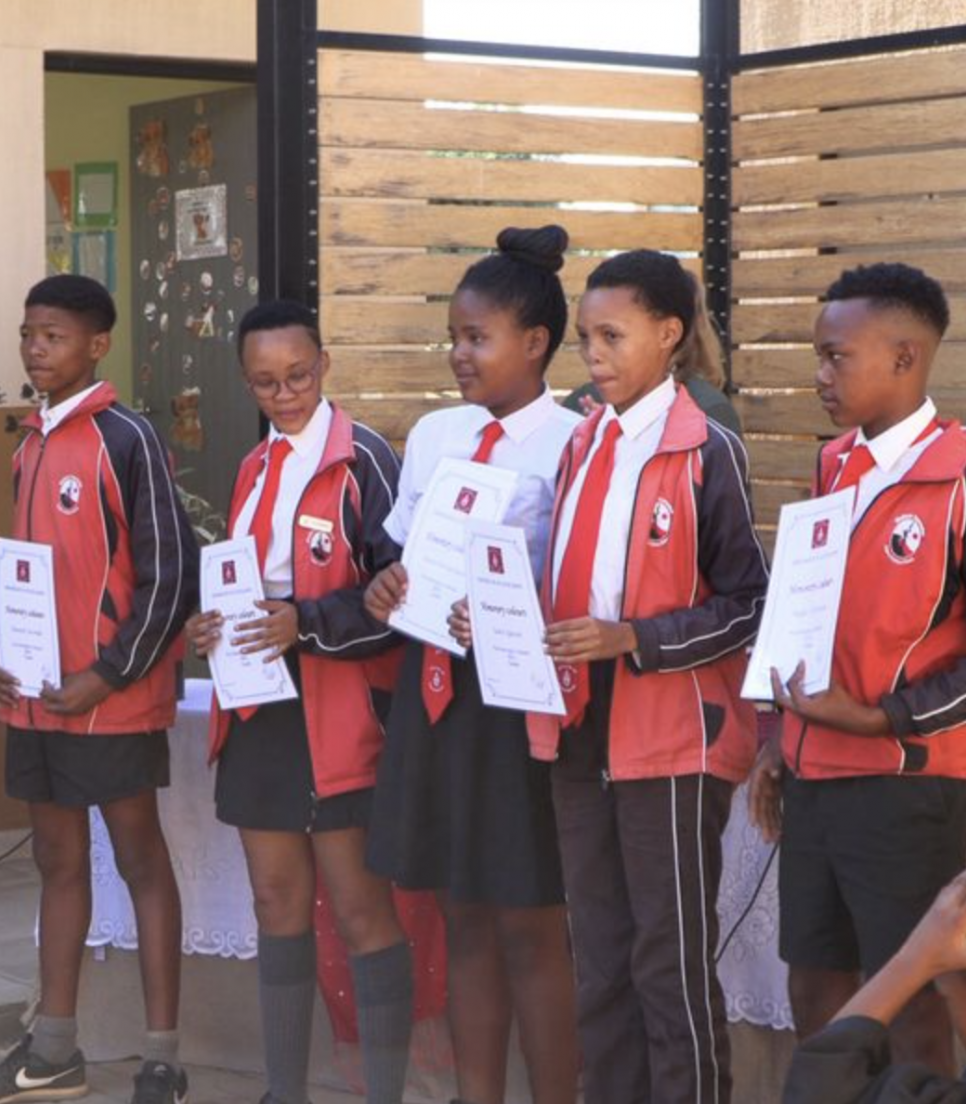 Cinq élèves San en uniforme reçoivent leurs certificats lors d’une cérémonie de fin d’année à l’école communautaire en Namibie, entourés de leurs enseignants.